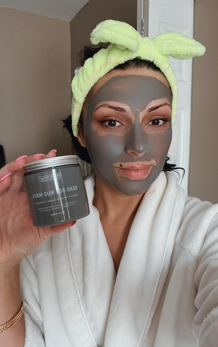 A reviewer in a bathrobe and hair wrap applying a dead sea mud mask, holding up the product jar.