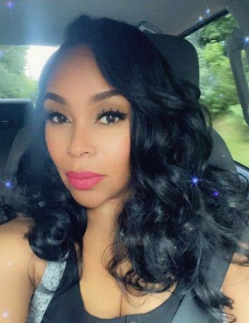 A reviewer with wavy hair sitting in a car, sporting bright lipstick and looking directly at the camera.