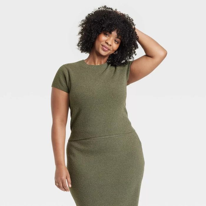 Woman in a form-fitting, short-sleeve dress confidently posing with one hand on her hip and the other playing with her curly hair. Shopping category image.
