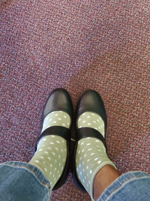 A reviewer stands on a carpeted floor wearing black Mary Jane shoes paired with polka-dot socks, showcasing a casual yet stylish look.