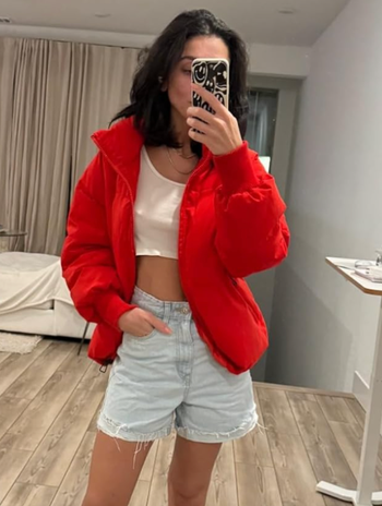 Person wearing a cropped white top, red jacket, and denim shorts takes a mirror selfie in a bedroom