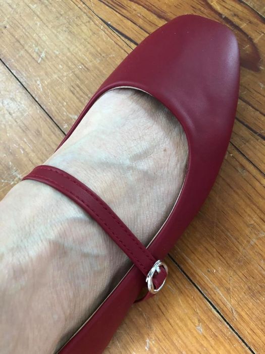 A close-up of a reviewer’s foot adorned with a red Mary Jane shoe featuring a strap and silver buckle, resting on a wooden floor.