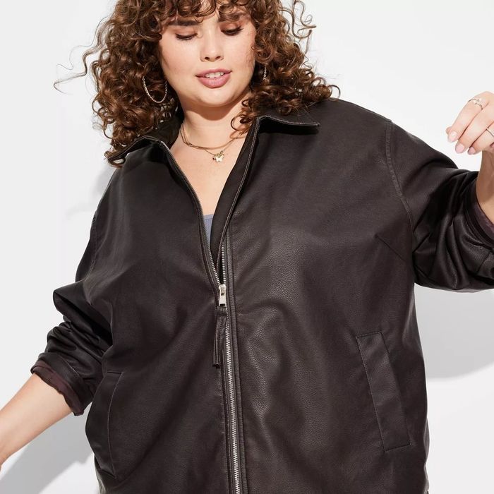 Person wearing a fashionable leather jacket, sporting curly hair and hoop earrings, gazing downward with an air of confidence.