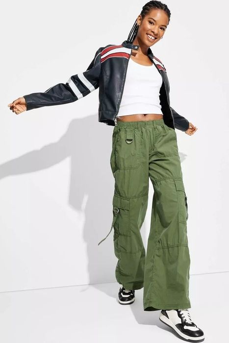 A beaming model strikes a pose in a white tank top, paired with a black-and-red-striped jacket and green cargo pants, finishing off the look with sneakers.