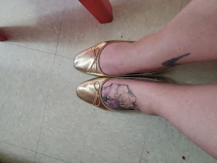A reviewer showcasing gold ballet flats, with a floral tattoo visible on their right foot. This image is featured in a Shopping article.