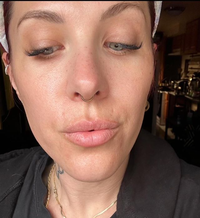 A close-up shot of an individual with a septum nose ring and long lashes, wearing a dark top and a white head wrap, seen indoors.
