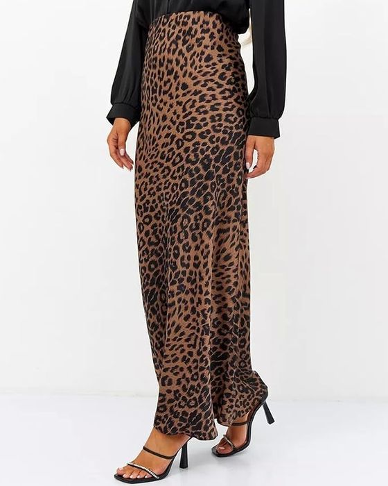A person in a black long-sleeve top, wearing a high-waisted leopard print maxi skirt and black high-heeled sandals. Only their lower body and hands are visible.
