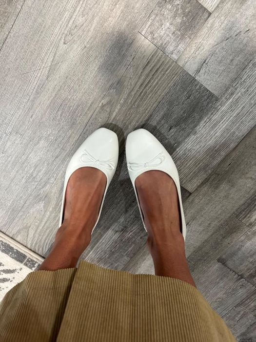 A reviewer sporting white flats with bows, standing on a wooden floor, visible from the knee down.