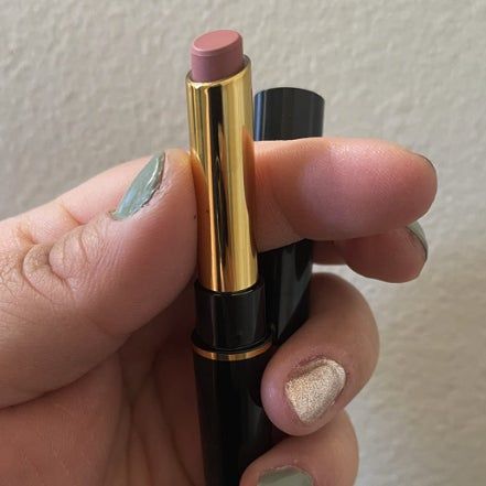 A close-up shot of a hand holding a black and gold lipstick tube featuring a soft pinkish nude shade.