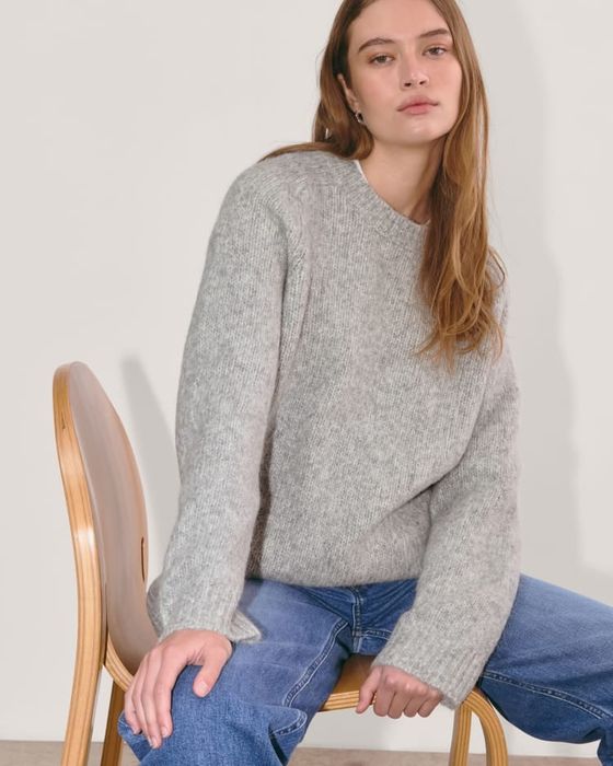 A woman sitting in a chair, wearing a cozy knit sweater and relaxed-fit jeans, featured in a shopping article.