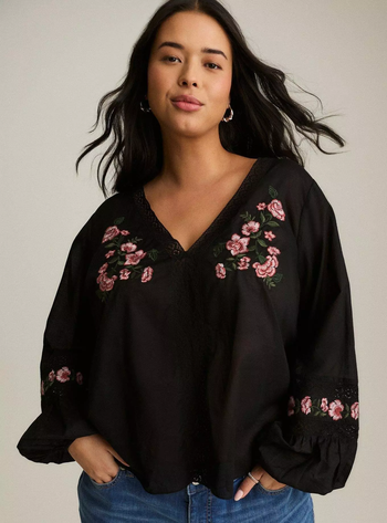 Model wearing a black blouse adorned with floral embroidery, striking a confident pose.