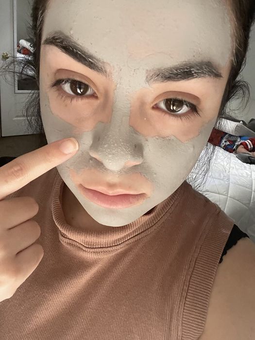 Someone applying a clay mask to their face, pointing to their cheek, in an indoor setting for a skincare shopping guide.