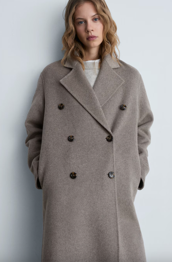A person wearing a long, double-breasted coat with a notched collar, standing against a neutral background.