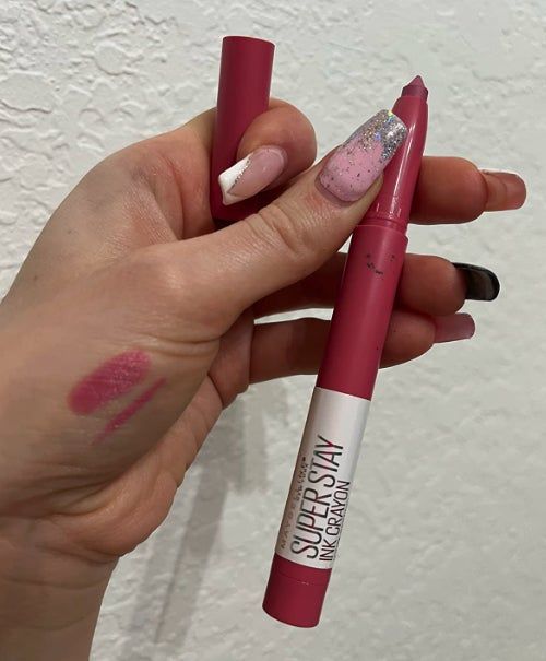 A hand with multicolored nails holds a SuperStay Ink Crayon lipstick, showcasing a swatch of the shade on the back of the hand.