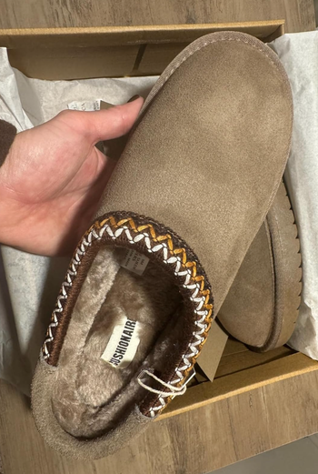 Hand holding a suede slipper with patterned trim in an open shoe box. The displayed item is categorized under Shopping