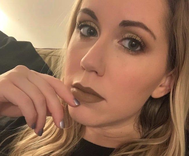 A reviewer with long blonde hair and a subtle makeup look, featuring brown lipstick and metallic eyeshadow, gazes into the camera while resting her chin on her hand.