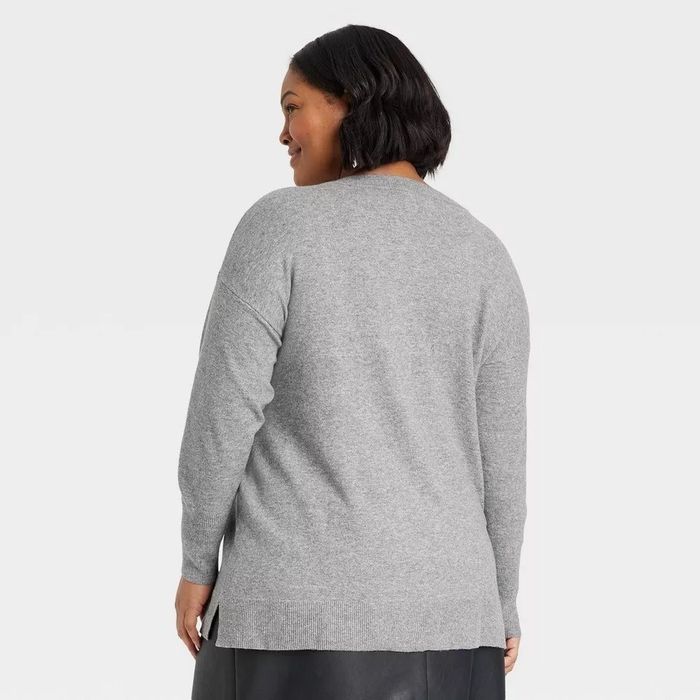 A person with short dark hair wears a loose-fitting gray sweater, turning slightly away from the camera to highlight the sweater’s stylish back design.