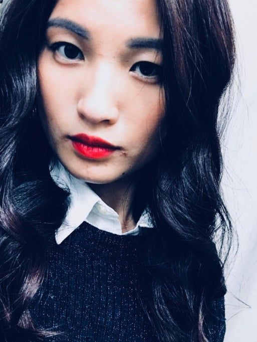 A reviewer with long, wavy hair and striking red lips is dressed in a black textured sweater over a white collared shirt, facing the camera with a confident expression.
