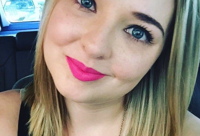 A reviewer with straight blonde hair grins at the camera, rocking vibrant pink lipstick.