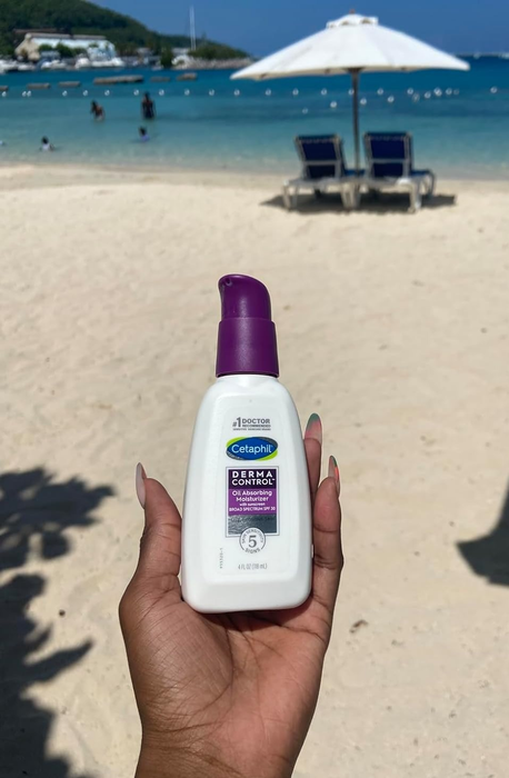 A reviewer holding a Cetaphil sunscreen bottle on a beach, with loungers, an umbrella, and the ocean in the background