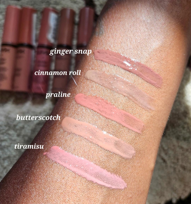 An arm with five liquid lipsticks swiped on it, each labeled with their shades: ginger snap, cinnamon roll, praline, butterscotch, and tiramisu, with matching tubes placed behind.