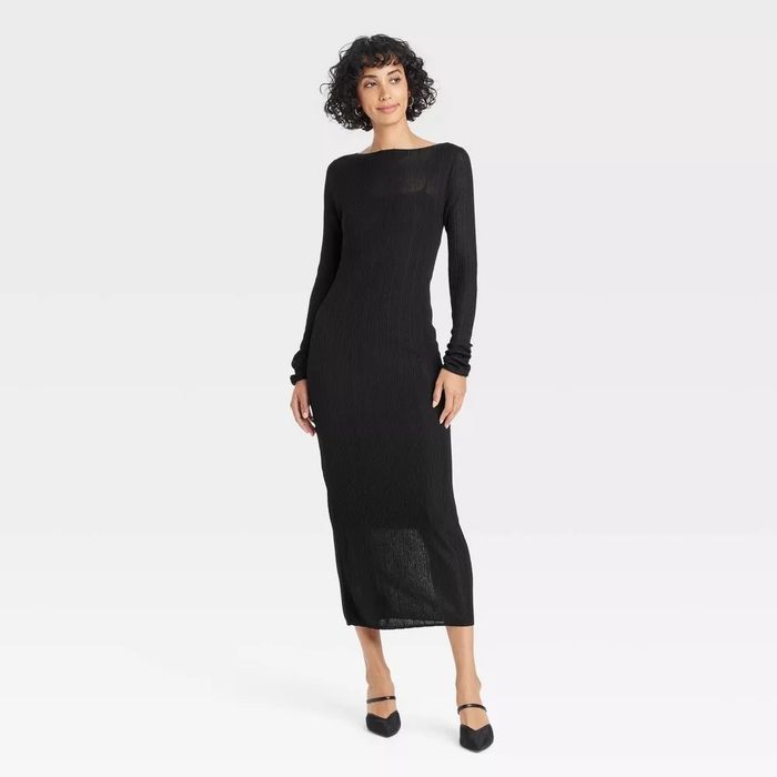 Individual wearing a long, figure-hugging black dress paired with black shoes, posed in a simple, neutral space.