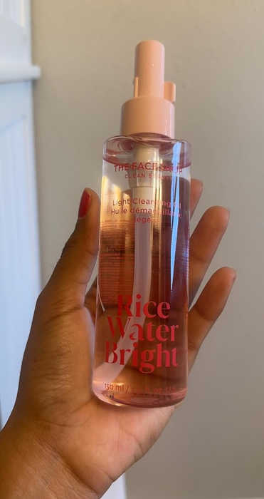 A reviewer holding a bottle of The Face Shop Rice Water Bright Light Cleansing Oil, featuring a spray nozzle and pink text on the label.