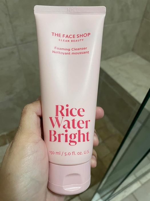 A reviewer holding a tube of The Face Shop Rice Water Bright Foaming Cleanser, with product details displayed in both English and French.