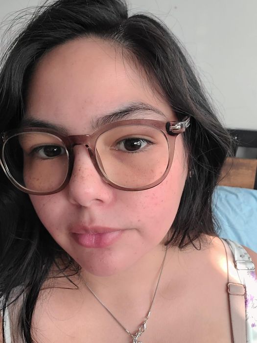 A woman with long dark hair and glasses stares at the camera in a close-up shot, wearing a floral top and a necklace.