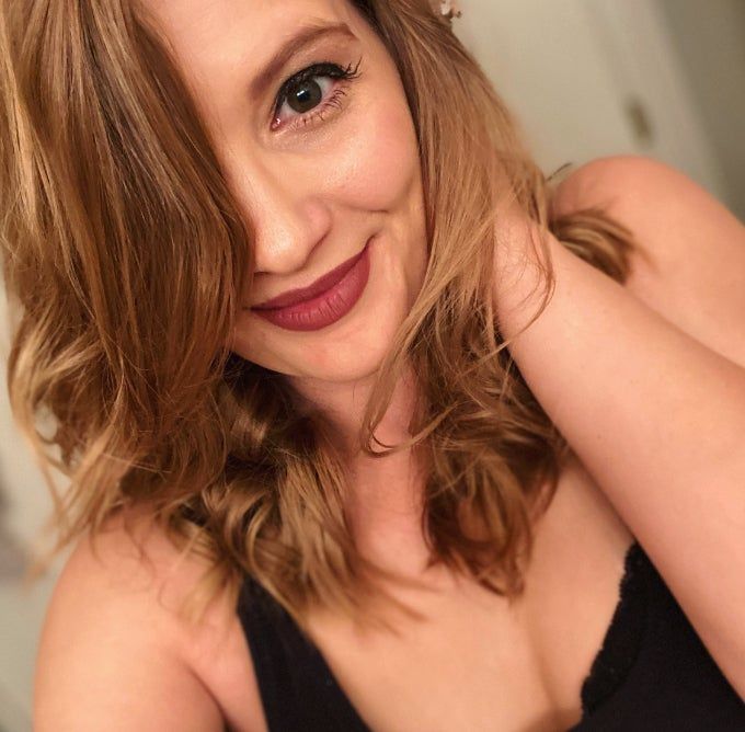 A person with wavy, shoulder-length hair smiles softly, wearing the Stila Stay All Day Lipstick.