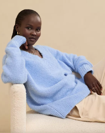 Person sitting casually, dressed in a cozy oversized sweater and light-colored pants, striking a relaxed pose. A trendy outfit featured in a shopping article.