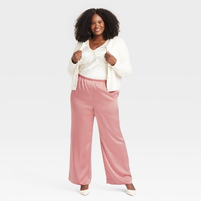 A woman confidently smiles, wearing a white cardigan and pink wide-leg pants, possibly modeling for a fashion campaign.
