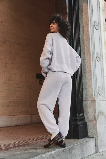 Model in a relaxed-fit sweater and matching pants, smiling and posing outside an urban building, featured in a shopping article.