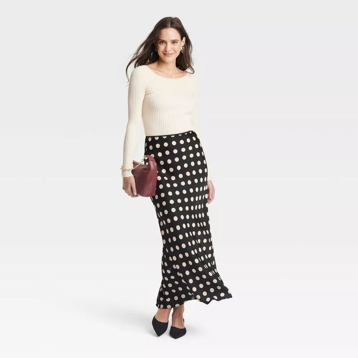 Model wearing a sleek long-sleeve top and a polka-dot maxi skirt, clutch in hand, posing against a plain backdrop.