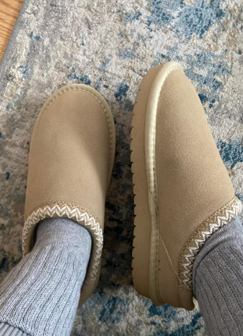 Feet wearing beige slippers with a knit trim resting on a patterned rug
