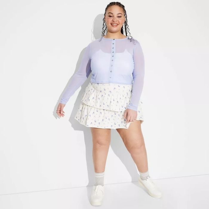 Person in a chic, sheer-sleeved blouse and floral tiered skirt, confidently posing in casual white sneakers.