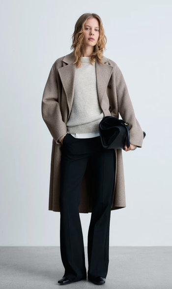 A person dressed in a long, tailored coat, sweater, and trousers, carrying a black handbag.