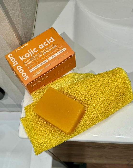 The kojic acid soap bar placed on a yellow mesh exfoliating cloth next to its product box. The soap is marketed for dark spot reduction and skin brightening.