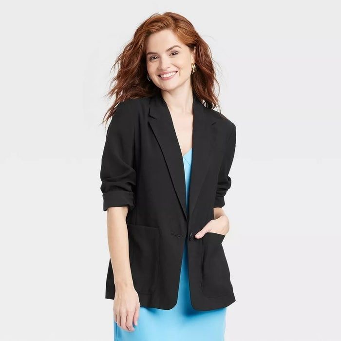 Person wearing a sleek black blazer over a blue dress, striking a confident pose and smiling.