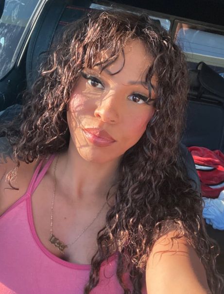 A reviewer with curly hair, wearing a pink top, sits in a car while sporting the shade called Androgyny.