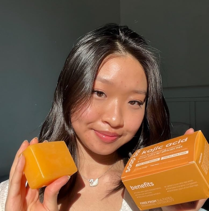 A reviewer holds the kojic acid soap bar, smiling at the camera, with the soap’s packaging highlighting its benefits.