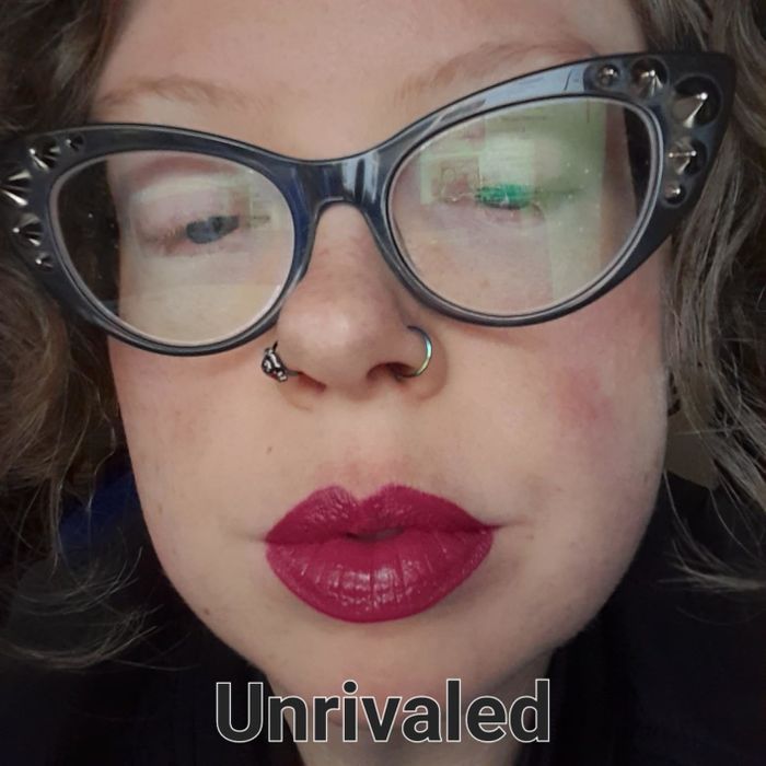 A close-up of a person sporting cat-eye glasses with intricate frames, a nose ring, and vibrant lipstick. Text at the bottom reads,