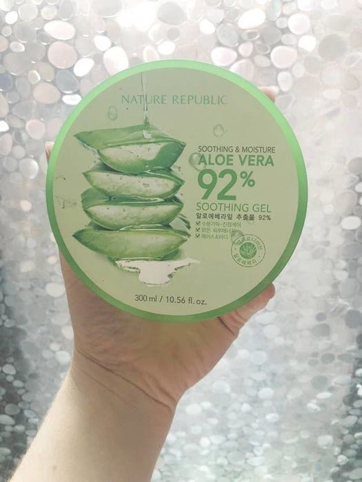 A hand holding the green tub of aloe vera gel in front of a window, showing its refreshing appeal.