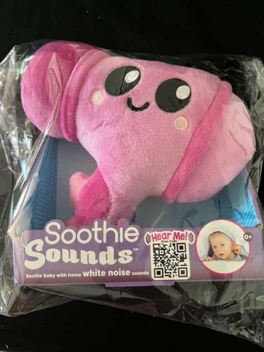 A Soothie Sounds plush toy wrapped in clear plastic, offering soothing home white noise sounds for babies. The packaging includes a baby image and a QR code for more details.