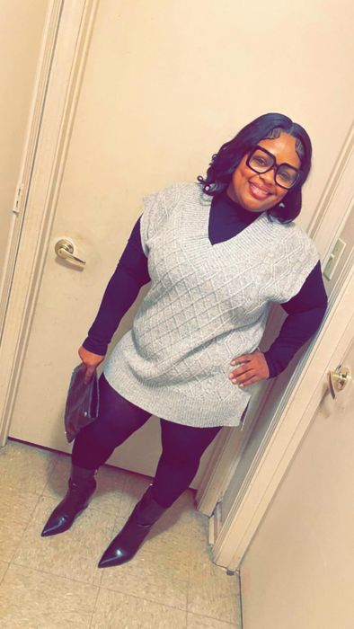 Person smiling while rocking black leggings, boots, a black turtleneck, and a textured gray vest. Holding a dark handbag, standing in a cozy indoor setting.