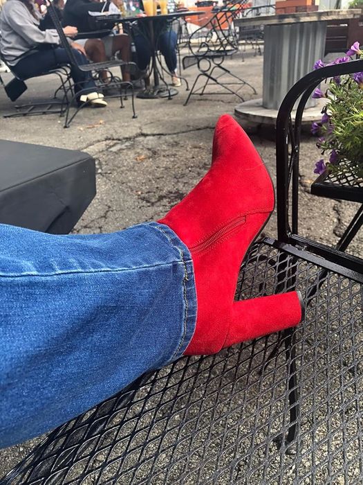 A reviewer dressed in red high-heeled boots and jeans, seated at an outdoor café surrounded by metal chairs and tables