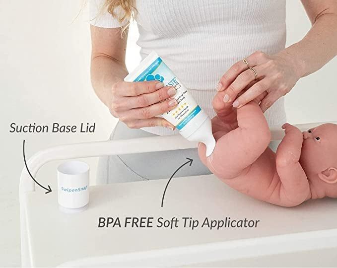 A person applying cream to their hand from a tube, next to a baby doll with a suction base lid and soft tip applicator