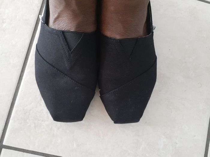 A reviewer sporting sleek black slip-on shoes on a tiled floor, showing off a clean, stylish look.
