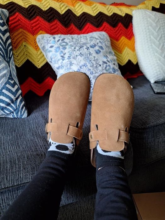 A reviewer wearing brown suede clogs over black socks, lounging on a couch surrounded by patterned pillows in the background.