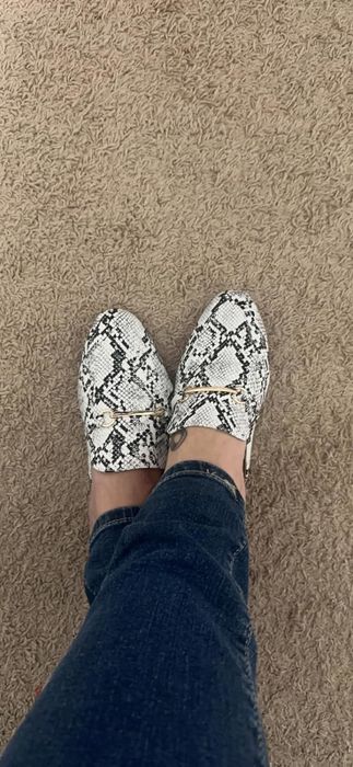 A reviewer wearing stylish snake print loafers paired with jeans, standing on a carpeted floor